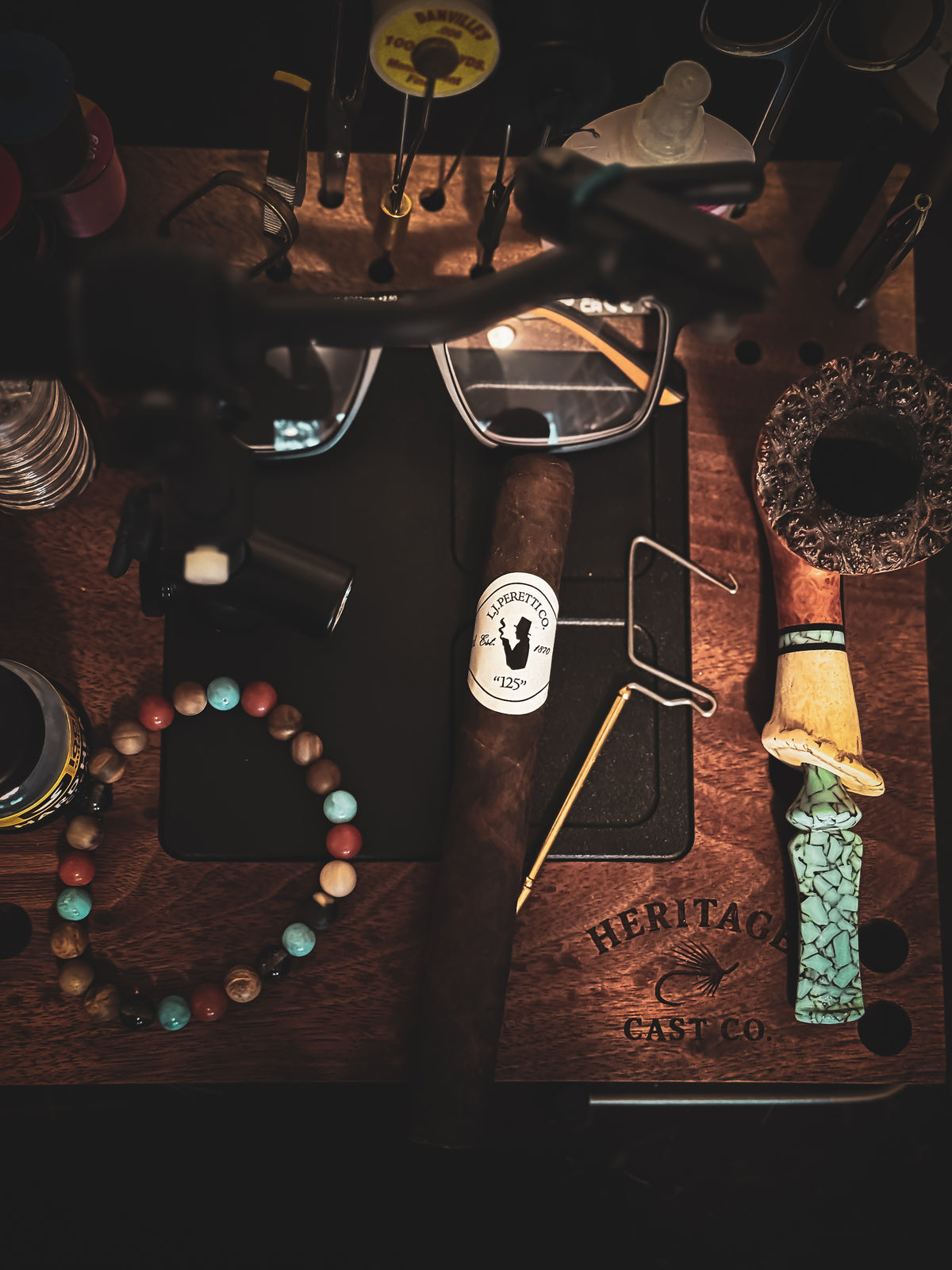 A top-down view of a wooden desk or tray holding a cigar, a beaded bracelet, eyeglasses, a smoking accessory, and a branded wooden plaque engraved with 'HERITAGE CASCO'.