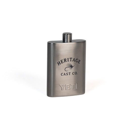 A brushed stainless steel hip flask with a black screw cap, featuring engraved logos for 'Heritage Cast Co.' and 'Yeti' on a clean white background.