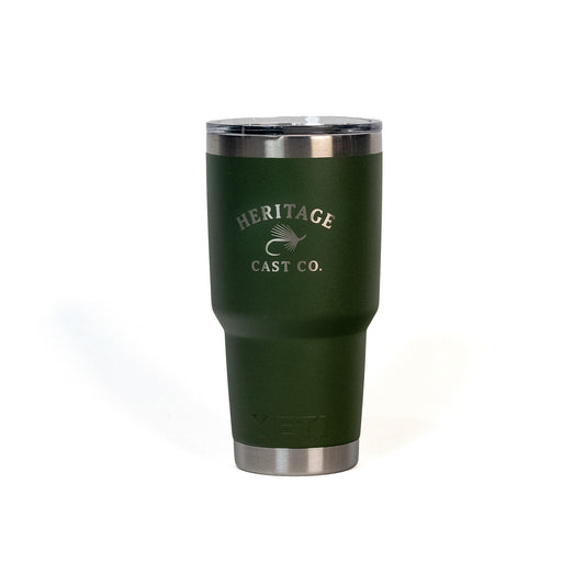 A dark green insulated travel tumbler with stainless steel accents and a clear lid, featuring the 'Heritage Cast Co.' logo printed on the front, set against a plain white background.
