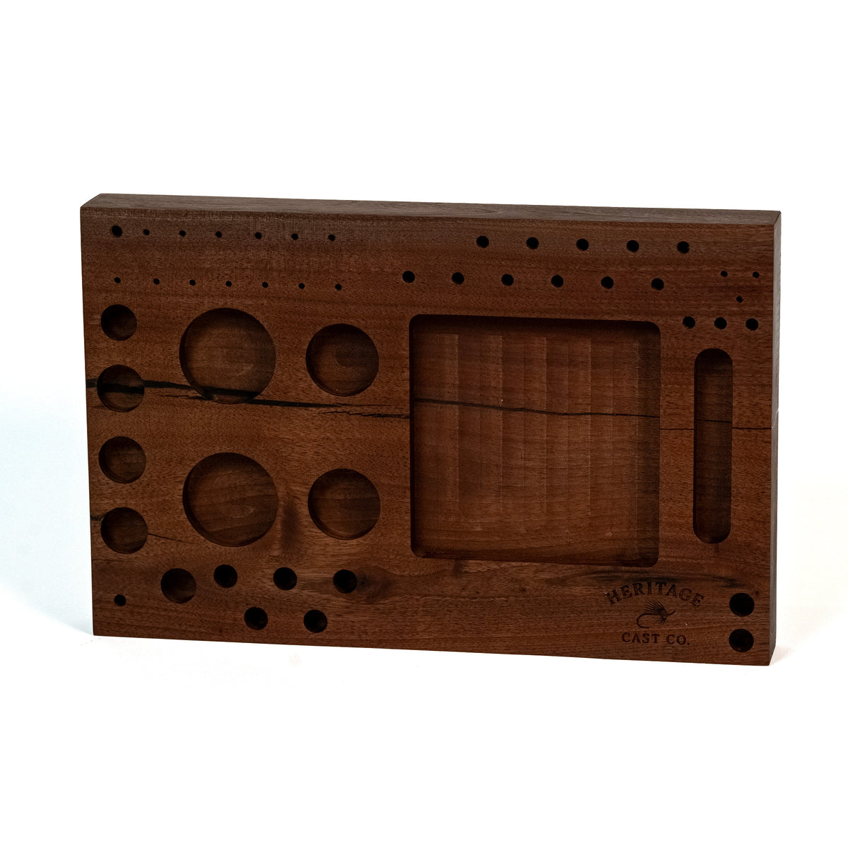 A rectangular dark brown wooden board with engraved circular and oval cutouts, a central recessed panel, and small dot markings across the top, photographed on a white background. The brand name 'Heritage Cast Co' is engraved in the lower right corner.