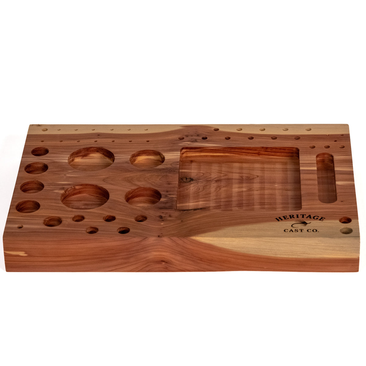 A rectangular wooden fly-tying station board with multiple circular and oval recesses, a large central slot, and engraved branding reading 'HERITAGE CAST CO' on a plain white background.