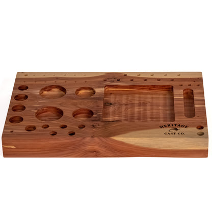 A rectangular wooden fly-tying station board with multiple circular and oval recesses, a large central slot, and engraved branding reading 'HERITAGE CAST CO' on a plain white background.