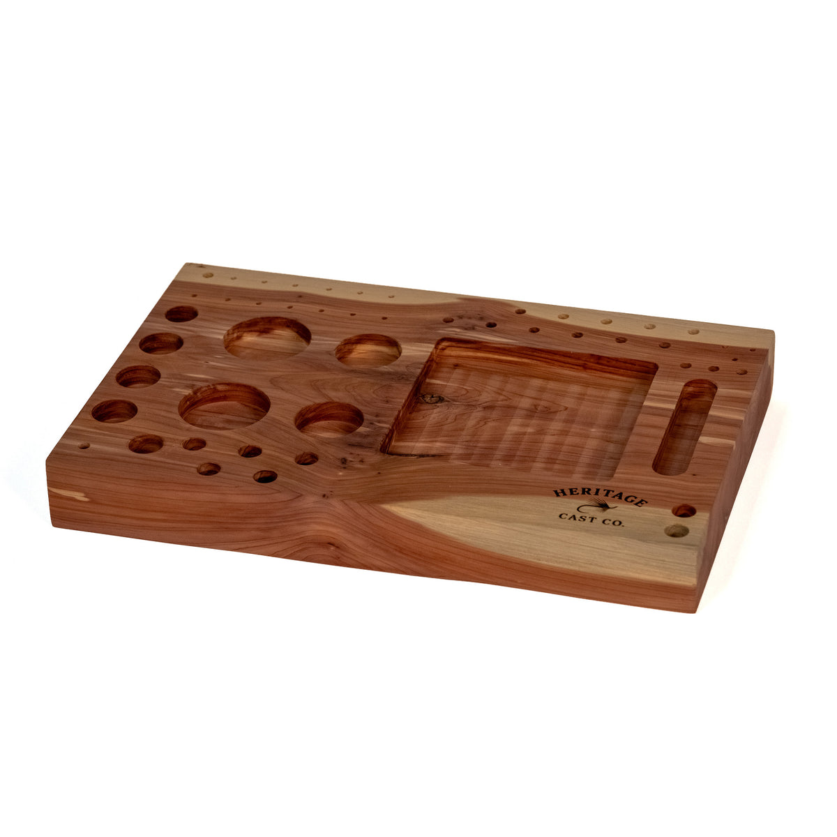 A wooden fly-tying tool set by Heritage Cast Co., featuring a polished wood block with multiple circular and oval holes, a recessed square section, and a vertical slot, shown on a white background.