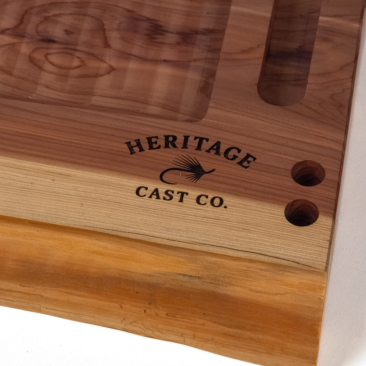 Close-up of a wooden fly-tying board or tray with a natural wood grain finish, featuring a cutout slot and two circular holes. The brand name 'HERITAGE CAST CO.